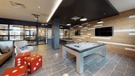 This single-floor amenity space combines contemporary design with a range of social and recreational features. The kitchenette runs along one wall, showcasing warm wood cabinetry, a striking black subway tile backsplash, white quartz countertops, and stainless steel appliances including a built-in microwave and undercounter refrigerator. A wall-mounted flat-screen TV is integrated seamlessly above the counter, adding a functional entertainment element to the space.

The main open area serves as both a gathering and game zone, anchored by a sleek pool table and a ping-pong table positioned beneath bold pendant lighting. A textured wood accent wall, adorned with a mounted television and decorative tile lettering, provides a warm visual contrast to the dark ceiling and structural columns. Oversized red dice add a playful design touch throughout.

A communal dining area with a long bar-height table and black stools flows naturally from the kitchenette toward glass-panel doors, which allow natural light to filter in and provide a visual connection to adjacent spaces. A darker lounge nook, featuring deep charcoal walls, slate tile flooring, and a swivel accent chair, offers a quieter retreat within the overall layout. The consistent use of slate-tone flooring throughout unifies the entire space.
