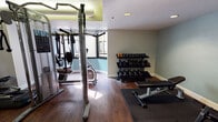 This fitness center is a comprehensively outfitted facility designed to support a wide range of workout routines. The space is finished with rich hardwood-style flooring throughout, paired with neutral-toned walls accented by a soft teal feature wall that creates a calm, focused atmosphere. Recessed and cove lighting illuminate the room evenly, complemented by a textured ceiling that adds a classic touch.

The cardio zone is well-stocked with multiple treadmills and an elliptical trainer, neatly arranged in a row and reflected by a large wall-to-wall mirror that visually expands the space and allows users to monitor their form. A wall-mounted flat-screen television is positioned for easy viewing during workouts.

The strength training area includes a multi-function cable machine offering a variety of resistance exercises, a stationary bike, and a dedicated free weights section featuring a full three-tier dumbbell rack with a wide range of weights. A sturdy adjustable bench and rubber floor mats provide a safe surface for floor exercises and stretching.

Additional amenities include what appears to be a compact refrigerator, a waste receptacle, and access to an adjoining room, likely a restroom or changing facility. Natural light filters in through a window, enhancing the overall brightness of the space. The layout is thoughtfully organized to maximize usability and comfort for all fitness levels.