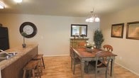 This single-story space showcases a warm and inviting dining area that combines rustic charm with classic design elements. The dining room centers around a solid wood dining table with a distressed finish, paired with elegantly carved chairs featuring decorative back detailing. Light hardwood flooring runs throughout, adding warmth and continuity to the space.

A chandelier with multiple globe lights hangs above the dining table, providing focused ambient lighting and serving as a tasteful focal point. A vintage-style sideboard in a muted sage green finish stands against the wall, topped with a framed mirror featuring a dark wood border, adding depth and visual interest to the room. Decorative accents, including potted greenery and a floral centerpiece on the table, enhance the natural, homey atmosphere.

A large round wall clock with a dark frame adds a classic decorative touch to one wall, while a partial view of a kitchen counter with bar stools suggests a connected, open-concept layout. Neutral beige walls complement the warm tones of the furnishings, creating a cohesive and comfortable environment throughout this single-level space.