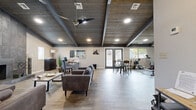 Located at 2029 N Woodlawn St in Wichita, Kansas, this single-floor fitness and community facility offers a thoughtfully designed space that balances active and social environments. The interior is defined by striking vaulted ceilings clad in rustic wood planking, accented by bold dark exposed beams that run the length of the space, lending a modern-industrial character throughout.

The exercise room is well-appointed with a variety of cardio equipment, including treadmills, an elliptical, and spin bikes, all set against light wood-look flooring. Natural light filters in through multiple windows, while a wall-mounted flat-screen TV adds functionality for workout sessions. A dedicated accent wall in muted teal provides a visual anchor to the fitness area.

Adjacent to the exercise zone, an open lounge and social area features comfortable seating arrangements, a bistro-style dining table, and a workstation, making it a versatile space for relaxation or casual gatherings. A stone fireplace column adds warmth and texture to the lounge. French doors open toward the exterior, enhancing the connection to the outdoors and allowing ample natural light. The entryway area includes additional seating and wall art, contributing to the space's welcoming, curated atmosphere. Recessed lighting and ceiling fans ensure the entire space remains bright and comfortable year-round.