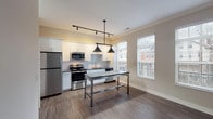 Located at 1221 E Broadway in Louisville's vibrant Highlands neighborhood, this single-floor unit showcases a thoughtfully designed, modern interior with consistent hardwood-style flooring and a neutral color palette throughout. The open kitchen is a standout feature, equipped with white shaker cabinetry, dark granite-style countertops, a classic subway tile backsplash, and a full suite of stainless steel appliances including a refrigerator, range, microwave, and dishwasher. A central island with a dark countertop provides additional workspace and is complemented by stylish industrial pendant lighting overhead. Track lighting adds functional ambiance to the cooking area, while large windows allow natural light to fill the space and offer views of the surrounding neighborhood. The bedroom is a spacious, light-filled room with crown molding and a distinctive dark-stained sliding barn door that opens to the en-suite bathroom. The bathroom is elegantly appointed with marble-look wall tile in the walk-in shower, wood-grain vinyl flooring, a white vanity with a speckled countertop, and recessed lighting, creating a clean and spa-like atmosphere. Together, these spaces form a cohesive, contemporary living environment with quality finishes throughout.