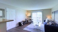 This single-floor space centers around a well-appointed living area that balances functionality with a clean, modern aesthetic. The room features dark hardwood-style flooring that contrasts attractively with the light neutral walls and white trim, creating a crisp, polished look throughout. Natural light is a defining characteristic of the space, streaming generously through a large sliding glass door dressed with vertical blinds, which also provides direct access to an outdoor area with visible greenery beyond — a pleasant extension of the living space.

The layout is open and accommodating, with ample room for a full seating arrangement. A low-profile media console serves as a practical storage and display surface along one wall, while a floor lamp adds warm ambient lighting to complement the natural light. The entry door is positioned conveniently within the living area, featuring a classic six-panel white design. A striped area rug in cool blue and green tones anchors the seating zone, adding a layer of color and texture to the overall aesthetic. The space presents a comfortable, move-in-ready atmosphere suitable for everyday living.