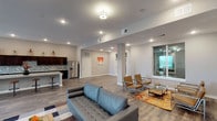 This contemporary single-floor amenity space is thoughtfully designed to foster comfort and community. The open-plan layout is anchored by rich hardwood-style flooring that flows seamlessly throughout, unifying the various functional zones within the space.

The kitchen area is a standout feature, showcasing an expansive granite-topped island with bar stool seating, dark espresso cabinetry, a mosaic tile backsplash, stainless steel appliances including a French door refrigerator, and ample counter space — ideal for gatherings and events.

The lounge areas are generously proportioned, offering multiple distinct seating configurations. A tufted gray sofa paired with a bold geometric area rug anchors one zone, while a separate arrangement of sleek tan leather chairs with chrome-framed glass tables provides a more intimate setting. Decorative wall art, including metallic sculptural pieces, adds a curated, modern aesthetic throughout.

Large windows along the exterior walls invite an abundance of natural light and offer views of the surrounding landscape, creating an airy, welcoming ambiance. Recessed lighting and flush-mount fixtures provide warm, even illumination throughout. Additional areas visible through open corridors suggest dedicated workspace or business center access, further enhancing the functionality of this well-appointed community space.