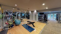 This fitness center is a comprehensively outfitted single-level facility designed to support a wide range of workout routines. The space is divided into distinct functional zones, each tailored to a specific type of exercise. The flooring transitions between patterned commercial carpet tiles in the cardio and strength areas and smooth hardwood in the dedicated stretching and floor exercise zone, providing appropriate surfaces for each activity.

The cardio section lines one side of the facility and features a row of treadmills and elliptical trainers by True Fitness, each equipped with individual display screens for performance tracking. A wall of floor-to-ceiling mirrors runs alongside this area, visually expanding the space and allowing users to monitor their form. A wall-mounted flat-screen television adds an entertainment option for those on the machines.

The strength training area houses a selection of cable and resistance machines, along with a rack of medicine balls and stability balls for functional training. The stretch zone features a padded workout mat, an adjustable bench, and open floor space. Recessed lighting throughout creates a clean, well-lit environment. French doors at one end open toward an outdoor pool area, bringing in natural light and a refreshing view.