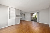 This single-floor residence offers a well-organized layout that balances open living areas with functional private spaces. The living room is a standout feature, showcasing rich, dark hardwood-style flooring and a ceiling fan overhead. A sliding glass door with vertical blinds opens to an outdoor patio framed by a distinctive arched entry, allowing natural light and garden views to fill the space. A side window with horizontal blinds adds further brightness.

The kitchen is positioned adjacent to the living area and features a peninsula with a breakfast bar, built-in open shelving, and white cabinetry. Stone-look countertops, a double-basin sink, and white appliances including a dishwasher and range complete the space. A window over the sink provides natural light.

The entry area transitions between the kitchen and a secondary living zone, with tile flooring at the threshold and wood-look flooring throughout. Built-in shelving near the entry adds practical storage.

The bedrooms are carpeted in neutral tones and receive natural light through windows fitted with blinds. The bathroom includes a full tub and shower combination with chrome fixtures. Textured ceilings are consistent throughout, contributing to the unit's cohesive aesthetic.