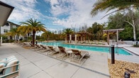 This single-level outdoor amenity space is designed with a resort-inspired aesthetic, offering a comprehensive range of leisure features set within a beautifully landscaped environment. At the heart of the space is a generously sized swimming pool with crystal-clear blue water, surrounded by a broad concrete sun deck lined with wood-and-white lounge chairs ideal for sunbathing and relaxation. Tall palm trees are strategically placed throughout the pool area, enhancing the tropical ambiance and providing natural shade.

Adjacent to the pool is a covered outdoor patio supported by white columns with stacked stone accents, creating an inviting and architecturally refined gathering area. This shaded space is furnished with plush sectional sofas and accent chairs arranged around a central fire pit, making it a versatile spot for both daytime lounging and evening socializing. A wall-mounted flat-screen television adds an entertainment element to the covered area.

A pergola structure with string lights is positioned at the far end of the pool deck, offering a charming and intimate setting for outdoor dining or relaxation. The building exterior features crisp white siding, dark-trimmed large-pane windows, and a dark shingled roof, complemented by thoughtfully designed landscaping with ornamental grasses, shrubs, and ground-level lighting that enhances the space after dark.
