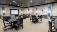This single-floor multipurpose room is designed to accommodate both entertainment and group gatherings with ease. At the focal point of the space is a large projection screen mounted on the front wall, paired with a ceiling-mounted projector, making it well-suited for film screenings, presentations, or events. The room is furnished with a thoughtful mix of seating options: plush reclining lounge chairs are positioned toward the screen for an immersive viewing experience, while several round tables with upholstered dining-style chairs are arranged throughout the space to support social gatherings or meetings. The neutral carpeted flooring and padded acoustic wall panels in a soft grey tone contribute to both the aesthetic and sound quality of the room. Teal accent panels frame the walls, adding a refined touch of color to the otherwise understated palette. Recessed ceiling lighting provides even, ambient illumination throughout, complemented by subtle wall sconces that enhance the room's warm atmosphere. A framed piece of artwork is displayed prominently on the back wall, adding a finishing decorative element. Overall, the space strikes a balance between functional versatility and comfortable, polished design.