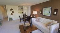 This single-floor residence showcases an open-concept layout that seamlessly connects the living, dining, and kitchen areas into one unified, flowing space. The living area is anchored by a crisp white sofa and a rich wooden coffee table adorned with decorative accents, set against plush neutral carpeting that runs throughout. A bold chocolate-brown accent wall adds depth and warmth to the living space, complemented by framed artwork and a wall-mounted sconce that contribute to the refined ambiance. A tall floor lamp provides layered lighting alongside the natural overhead illumination.

The dining area transitions naturally from the living space, featuring a glass-top table surrounded by dark leather chairs, creating an elegant yet functional setting. A pendant light hangs above, drawing the eye upward and defining the dining zone within the open plan.

The kitchen, visible in the background, offers white cabinetry, a distinctive wine rack built into the upper storage, and under-cabinet lighting that highlights the workspace. The overall design is cohesive, with a warm neutral palette, thoughtful lighting, and carefully chosen furnishings that give the space a polished, contemporary character.