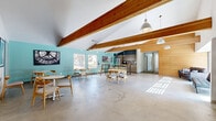 Located at 1200 S Oneida St in Denver, CO, this single-story community or amenity space offers a thoughtfully designed interior that blends modern aesthetics with warm, natural materials. The main gathering area commands attention with dramatically vaulted ceilings supported by exposed natural wood beams, polished concrete floors, and an abundance of natural light streaming through large windows and floor-to-ceiling sliding glass doors. Vibrant teal-painted walls, pendant lighting, and track lighting add energy and character to the open layout, which features multiple seating configurations including communal dining tables, high-top bar seating, and casual lounge areas. A kitchenette with cabinetry, a stainless steel refrigerator, and counter seating provides functional support for gatherings. A secondary lounge area offers a more relaxed atmosphere, accented by bold red and warm wood ceiling panels, a built-in window bench seat with views to the outdoors, and carpeted flooring for comfort. Additional areas include a quieter lounge with a rustic wood accent wall and glass-partitioned rooms for semi-private use. Red built-in shelving and accent walls throughout add cohesive pops of color, while restroom facilities and multiple access points ensure practicality across the entire space.