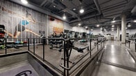 This expansive, single-floor fitness facility offers a generously sized workout environment thoughtfully divided into distinct training zones. The open-plan layout is anchored by a central raised platform with metal railing, providing clear sightlines across the entire floor and creating a natural separation between functional training areas.

The cardio zone houses an extensive row of treadmills, elliptical machines, steppers, and rowing machines, all equipped with individual digital displays. Mounted televisions are positioned throughout for member entertainment. A dedicated free weights area features a full rack of dumbbells alongside flat and adjustable benches, complemented by wall-to-wall mirrors that amplify the sense of space and aid in form monitoring.

The strength training section is furnished with a wide selection of cable machines, plate-loaded equipment, and multi-functional units, catering to a variety of muscle groups and fitness levels. Rubberised flooring covers the training areas for safety and durability, transitioning to polished concrete along the central walkway.

Exposed ductwork and structural columns contribute to an industrial aesthetic, while overhead lighting ensures the space is bright and functional throughout. Motivational typographic wall murals bearing words such as "Passion," "Focus," and "Succeed" reinforce the energetic ambiance. Changing facilities are accessible on-site.