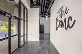 The front entrance of The Banc delivers a striking first impression through a carefully curated blend of industrial and contemporary design elements. Floor-to-ceiling black-framed glass doors and windows dominate the entry facade, flooding the space with natural light while offering clear views of the surrounding streetscape and mature trees outside. The polished metallic concrete floors extend throughout, featuring a dynamic swirled grey finish that adds visual depth and reflects light throughout the space. Crisp white walls serve as a clean canvas, most notably showcasing a large-scale brushscript "The Banc" wall graphic that immediately establishes brand identity upon entry. The soaring ceiling height enhances the sense of openness, with exposed ductwork and mechanical systems contributing to the industrial aesthetic. Bold navy blue trim and door surrounds provide rich color contrast against the white walls. A vibrant, large-format artwork depicting a headphone-wearing primate adds an eclectic, artistic touch to the corridor leading further into the space. A glimpse into an adjacent utility room reveals professional-grade equipment. Overall, the entrance conveys a polished, modern commercial environment with strong branding and deliberate design details.