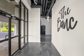 The front entrance of The Banc delivers a striking first impression through a carefully curated blend of industrial and contemporary design elements. Floor-to-ceiling black-framed glass doors and windows dominate the entry facade, flooding the space with natural light while offering clear views of the surrounding streetscape and mature trees outside. The polished metallic concrete floors extend throughout, featuring a dynamic swirled grey finish that adds visual depth and reflects light throughout the space. Crisp white walls serve as a clean canvas, most notably showcasing a large-scale brushscript "The Banc" wall graphic that immediately establishes brand identity upon entry. The soaring ceiling height enhances the sense of openness, with exposed ductwork and mechanical systems contributing to the industrial aesthetic. Bold navy blue trim and door surrounds provide rich color contrast against the white walls. A vibrant, large-format artwork depicting a headphone-wearing primate adds an eclectic, artistic touch to the corridor leading further into the space. A glimpse into an adjacent utility room reveals professional-grade equipment. Overall, the entrance conveys a polished, modern commercial environment with strong branding and deliberate design details.