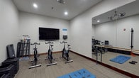 This single-floor fitness studio is thoughtfully designed to support a variety of exercise disciplines in a clean, functional environment. The room is finished with neutral white walls, dark wood baseboards, and low-pile carpeted flooring that provides both comfort and durability underfoot. Recessed ceiling lighting ensures the space is well-lit throughout.

The studio is equipped with multiple stationary cycling bikes arranged facing a wall-mounted flat-screen television, making it well-suited for guided cycling classes or independent cardio sessions. A small control panel or display unit is mounted beneath the TV for added convenience. A yoga mat on the floor further signals the room's versatility for stretching and floor-based exercises.

A full-length wall mirror spans a significant portion of one side of the room, creating a sense of expanded space while also serving a practical function for form-checking during workouts. Mounted in front of the mirror is a horizontal ballet barre, adding a dedicated zone for barre exercises, stretching, and balance training.

Overall, the studio offers a compact yet well-equipped environment for cardiovascular training, barre work, and flexibility routines, making it a versatile amenity for fitness enthusiasts.