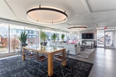 Located at 1810 Highpoint Ave in Richmond, VA, this single-floor amenity space offers a thoughtfully designed communal environment that balances social, recreational, and professional needs. The open layout is anchored by wide-plank hardwood-style flooring and crisp white ceilings with geometric tray detailing and circular recessed lighting fixtures that add architectural interest overhead.

The main lounge area features generous seating arrangements, including a large sectional sofa positioned beneath a dramatic circular ceiling cove, mid-century-inspired leather armchairs, and area rugs that define each zone. Floor-to-ceiling windows wrap the perimeter, flooding the space with abundant natural light and providing sweeping views of the surrounding Richmond streetscape and outdoor terrace.

A stylish kitchenette island with dark stone countertops, a built-in refrigerator, and coffee station is elegantly separated from the lounge by a striking vertical wood slat partition — a standout architectural feature that adds warmth and visual depth throughout the space.

The recreational zone includes a full-size billiards table and a wall-mounted television, making it well-suited for casual gatherings. A dedicated business center offers workstations set against a lush living green wall, paired with a round marble-top conference table for collaborative work. The overall ambiance is modern yet inviting, blending bold design elements with comfortable, functional furnishings.