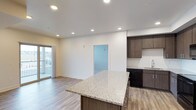 This single-floor unit showcases a thoughtfully designed open-concept layout that seamlessly connects the kitchen and living areas. The kitchen is a standout feature, equipped with dark wood-toned cabinetry that extends to the upper walls, providing generous storage. A large center island with a speckled granite countertop serves as both a workspace and casual gathering point, complemented by matching granite along the perimeter counters. Black appliances, including a refrigerator, range, microwave, and dishwasher, are integrated alongside a subway tile backsplash that adds a clean, contemporary touch.

Recessed lighting throughout the space ensures consistent, even illumination across the open floor plan. Light wood-toned luxury vinyl plank flooring runs continuously through the kitchen and living area, contributing to a cohesive and modern aesthetic. The living area benefits from natural light provided by a sliding glass door that opens to a private balcony with a railing, offering outdoor views.

A half-wall partition with a pass-through window separates an adjacent secondary space, adding architectural interest. Additional doors lead to other areas of the unit. The overall ambiance is clean, bright, and move-in ready with a contemporary design sensibility.