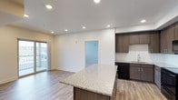 This single-floor unit showcases a thoughtfully designed open-concept layout that seamlessly connects the kitchen and living areas. The kitchen is a standout feature, equipped with dark wood-toned cabinetry that extends to the upper walls, providing generous storage. A large center island with a speckled granite countertop serves as both a workspace and casual gathering point, complemented by matching granite along the perimeter counters. Black appliances, including a refrigerator, range, microwave, and dishwasher, are integrated alongside a subway tile backsplash that adds a clean, contemporary touch.

Recessed lighting throughout the space ensures consistent, even illumination across the open floor plan. Light wood-toned luxury vinyl plank flooring runs continuously through the kitchen and living area, contributing to a cohesive and modern aesthetic. The living area benefits from natural light provided by a sliding glass door that opens to a private balcony with a railing, offering outdoor views.

A half-wall partition with a pass-through window separates an adjacent secondary space, adding architectural interest. Additional doors lead to other areas of the unit. The overall ambiance is clean, bright, and move-in ready with a contemporary design sensibility.