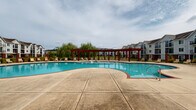 Located at 136 Crimson Rose Dr in Huntsville, Alabama, this two-story residential community features a standout outdoor amenity area centered around a generously sized freeform swimming pool. The pool's curvilinear design creates an inviting, resort-like atmosphere, with vivid blue water and a broad concrete pool deck that accommodates an abundance of lounge chairs for sunbathing and relaxation.

A striking deep-red pergola structure serves as the architectural focal point of the pool area, providing shaded gathering space along the poolside. The pergola's bold color contrasts attractively against the neutral tones of the surrounding buildings and landscape. Mature weeping willow trees frame the perimeter, adding a natural, serene quality to the outdoor setting.

The pool deck is enclosed by a classic black iron fence, offering both security and unobstructed views of the surrounding green lawn areas. A dedicated clubhouse building with red brick accents is visible nearby, complementing the overall community aesthetic. Ample lounge seating is arranged throughout the deck, providing residents with multiple areas to relax outdoors.

The multi-story residential buildings, featuring arched windows and neutral siding with brown rooflines, rise above the pool area and overlook the amenity space. Together, these elements create a well-appointed outdoor environment designed for leisure and community living.