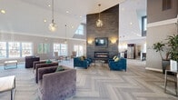 Located at 71 Mayfair Square in Danbury, CT, this thoughtfully designed single-floor community clubhouse offers an expansive and elegantly appointed gathering space. The centerpiece of the layout is a dramatic living area distinguished by soaring vaulted ceilings, a floor-to-ceiling dark tile accent wall, and a sleek linear gas fireplace topped by a wall-mounted television flanked by contemporary sconce lighting. Geometric pendant chandeliers hang overhead, casting warm light across herringbone-patterned carpeted floors that flow throughout the space. Multiple seating arrangements featuring plush velvet sofas and armchairs in rich teal and charcoal tones invite relaxed socializing, while large windows bathe the area in abundant natural light. Vibrant teal abstract artwork adds pops of color to the neutral gray walls. A dedicated lounge area offers a quieter retreat with additional seating and curated artwork. The kitchen is well-equipped with dark espresso cabinetry, stainless steel appliances including a full-size refrigerator, microwave, and dishwasher, complemented by geometric pendant lighting. A billiards table in the game area adds a recreational dimension. The dining room features patterned wallcovering, a glass-top table with upholstered seating, built-in bookshelves, and bright natural light. A glass-enclosed office or leasing area with a reception desk completes this refined and multifunctional space.