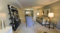 This single-floor residence offers a seamlessly connected living and dining layout, designed with both comfort and style in mind. The living area is anchored by plush, neutral-toned carpeting and features a contemporary seating arrangement, including a statement blue accent chair, a cushioned sofa with coordinating throw pillows, and a sleek media console with open shelving. A patterned area rug defines the space and adds visual texture underfoot.

A warm, sage-green accent wall frames the dining area in the background, where a round table is complemented by an elegant chandelier overhead, creating an intimate setting for meals. Natural light filters throughout, enhanced by a well-placed floor lamp that adds a soft, ambient glow to the room.

Decorative mirrors and framed artwork adorn the walls, contributing to a polished, curated aesthetic. The open sightlines between the living and dining zones promote a sense of spaciousness and ease of movement throughout the floor plan. The overall ambiance is warm and sophisticated, blending neutral tones with carefully chosen accent colors to create a cohesive and welcoming interior environment suited for both everyday living and entertaining.