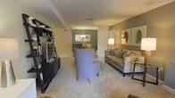 This single-floor residence offers a seamlessly connected living and dining layout, designed with both comfort and style in mind. The living area is anchored by plush, neutral-toned carpeting and features a contemporary seating arrangement, including a statement blue accent chair, a cushioned sofa with coordinating throw pillows, and a sleek media console with open shelving. A patterned area rug defines the space and adds visual texture underfoot.

A warm, sage-green accent wall frames the dining area in the background, where a round table is complemented by an elegant chandelier overhead, creating an intimate setting for meals. Natural light filters throughout, enhanced by a well-placed floor lamp that adds a soft, ambient glow to the room.

Decorative mirrors and framed artwork adorn the walls, contributing to a polished, curated aesthetic. The open sightlines between the living and dining zones promote a sense of spaciousness and ease of movement throughout the floor plan. The overall ambiance is warm and sophisticated, blending neutral tones with carefully chosen accent colors to create a cohesive and welcoming interior environment suited for both everyday living and entertaining.