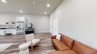 Located at 693 Morsani Pl in Tontitown, Arkansas, this single-floor space offers a well-designed open-concept layout that seamlessly integrates a kitchen, dining room, and living area. The interior is finished with warm-toned wood-look flooring throughout, neutral gray walls, and recessed lighting that ensures consistent, even illumination across the entire space.

The kitchen features crisp white shaker-style cabinetry paired with a striking dark granite countertop with bold white veining, providing a dramatic contrast. A stainless steel sink, an open display shelf above the counter, and a full-size stainless steel refrigerator complete the functional kitchen setup. Additional appliances, including a range, are also present.

The dining area is anchored by a large rectangular wood-tone table surrounded by upholstered chairs, set atop a patterned area rug. A standout architectural detail — an exposed wood beam accent — adds rustic warmth to the otherwise modern interior. Natural light enters through a window offering views of the surrounding landscape.

The living area features a cognac leather sectional sofa alongside a modern accent chair and side table, creating a relaxed yet stylish seating arrangement. A set of double white panel doors provides additional storage or utility access. The vaulted ceiling in the living zone adds a sense of openness and height to the space.