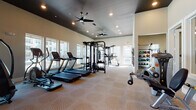This modern fitness center is a comprehensive, single-floor facility designed to support a full range of workout routines. The main exercise floor features patterned, low-pile carpeting and is anchored by a row of cardio machines, including treadmills, ellipticals, and a stair climber, all positioned along large windows that allow abundant natural light and offer views of an outdoor pool and surrounding building complex. A multi-station cable machine and additional strength training equipment occupy the center of the room, providing options for a wide variety of resistance exercises. A dedicated wall-mounted weight rack organizes an array of medicine balls, dumbbells, and kettlebells, keeping free weights neatly accessible. A separate studio space is distinguished by its bold, geometric carpet pattern in gold and gray tones, and houses spin bikes and exercise mats for group or individual training sessions. Full-length mirrors are strategically placed throughout to allow form monitoring during workouts. The dark-painted ceiling is fitted with recessed lighting and ceiling fans, maintaining both a stylish ambiance and comfortable airflow. A wall-mounted TV provides entertainment, and a stainless-steel sanitation station supports cleanliness throughout the facility.