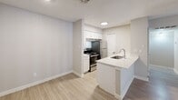 Located at 3303 S 2nd St in Austin, TX, this single-floor unit offers a clean, modern aesthetic with a neutral color palette and light wood-style flooring that runs consistently throughout the space. The open-concept layout connects the kitchen directly to the living area, promoting a seamless flow ideal for everyday living and entertaining. The kitchen is outfitted with white shaker-style cabinetry, a peninsula island with an undermount sink, a gas range, and a stainless steel refrigerator, all complemented by white countertops. The living area is generously sized, featuring a front entry door flanked by a window that allows natural light to filter in. A sliding glass door in the main living space opens to an outdoor area and is fitted with vertical blinds, while a ceiling fan overhead aids in air circulation. The bedroom continues the light flooring theme and includes a ceiling fan with light fixture and a window dressed with blinds. The bathroom features a tiled tub-and-shower combination with chrome fixtures, wood-look tile flooring, and clean white finishes throughout. Textured ceilings are present across the unit. The overall ambiance is bright, functional, and move-in ready.