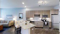This single-floor residence at Overture offers a thoughtfully designed open-concept layout that seamlessly blends style and functionality. The kitchen serves as a focal point of the space, showcasing light wood-toned cabinetry that provides ample storage alongside stainless steel appliances, including a range, microwave, and dishwasher. A striking quartz-topped island anchors the kitchen, offering additional prep space and seating for casual dining with upholstered bar stools. Under-cabinet lighting illuminates the tasteful mosaic tile backsplash, adding texture and visual interest to the cooking area. The kitchen flows naturally into the adjacent living area, creating a connected, social atmosphere ideal for entertaining or everyday living. Wide-plank wood-style flooring runs throughout, grounding the space with warmth and durability. Soft, neutral wall tones complement the contemporary finishes, while recessed and track lighting provide versatile illumination throughout. The overall design balances modern aesthetics with practical, livable comfort.