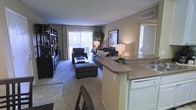 This single-floor residence is designed around an open-concept floor plan that encourages a natural flow between the kitchen and living areas. The kitchen features a wraparound breakfast bar with a neutral countertop, white cabinetry, and a dishwasher, making it both functional and visually connected to the rest of the space. A dining area sits adjacent, providing a transitional zone between the two main spaces.

The living room is carpeted and generously proportioned, furnished with a sofa, armchair, and ottoman arranged around a glass-topped coffee table. A tall bookshelf unit along one wall offers ample storage and display space, while framed wall art adds a decorative touch. The room is brightened by a large sliding glass door that opens to a private outdoor area, flanked by curtain panels and surrounded by natural greenery visible through the glass. Warm ambient lighting from table lamps complements the natural light throughout the day.

Neutral wall tones and warm-toned accents contribute to a cohesive, welcoming atmosphere throughout the space.