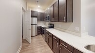 This thoughtfully designed multi-level residence offers a clean, contemporary aesthetic throughout its well-proportioned living spaces. The kitchen is a functional highlight, featuring rich dark-stained cabinetry with brushed chrome hardware, bright white countertops, and a full suite of stainless steel appliances including a refrigerator, range, and microwave. Light wood-look flooring runs through the kitchen, adding warmth and ease of maintenance to the space.

The main living area is generously sized, finished with neutral wall-to-wall carpeting that adds comfort underfoot. A sliding glass door opens directly to a private balcony, allowing natural light to fill the room and providing views of the surrounding neighborhood. Recessed lighting overhead ensures the space remains bright throughout the day.

An open upper landing area connects the living spaces and stairwell, carpeted to match the rest of the upper level. The staircase itself is fitted with plush carpet and a sleek metal handrail along one wall, providing a safe and stylish transition between levels.

The full bathroom is finished with light wood-look flooring, a white tub-shower combination, a toilet, and a vanity with dark wood cabinetry topped with a white countertop and a mirror above. The overall palette of neutral tones and consistent finishes throughout the space creates a cohesive, move-in-ready environment.