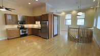 This two-story space offers a thoughtfully designed layout that combines functionality with classic style. The main level showcases an open-concept floor plan where the kitchen flows seamlessly into the living area, all unified by warm, light-toned hardwood floors that extend throughout.

The kitchen is well-appointed with rich wood cabinetry, stainless steel appliances including a refrigerator, range, and microwave, as well as a double sink set beneath a window. A central kitchen island adds additional workspace and helps define the cooking area within the open layout.

The living space beyond the kitchen is bright and airy, benefiting from multiple windows that invite natural light deep into the interior. Recessed lighting and a ceiling fan complement the natural illumination throughout.

A standout architectural feature of the space is the elegantly curved staircase with a classic wooden banister, adding a refined aesthetic detail that connects the two levels. The curved railing adds a sense of character and craftsmanship to the overall design.

Crown molding details and clean wall finishes contribute to a polished, well-maintained ambiance throughout. The combination of open living spaces, quality finishes, and a multi-level layout makes this a versatile and inviting space.