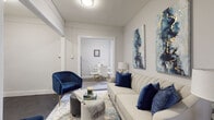 This well-appointed single-floor residence offers a cohesive blend of classic architectural character and modern finishes throughout its thoughtfully laid-out spaces. The living room serves as the heart of the home, featuring dark hardwood flooring, neutral walls, and curated décor accented with deep navy blue tones. Bold abstract artwork adorns the walls, complementing the elegant velvet seating and patterned area rug that anchor the space. An open doorway connects the living area to a separate dining room, where natural light filters in through large windows framed in dark trim, highlighting a radiator that speaks to the building's classic heritage. The kitchen is a standout feature, boasting rich, dark espresso cabinetry, white quartz countertops, a crisp subway tile backsplash, and stainless steel appliances including a gas range, all set atop light wood-tone flooring. Two bedrooms offer comfortable retreats; the primary bedroom is brightened by corner windows and features dark hardwood floors, while the second bedroom provides a serene, minimalist environment. The bathroom is elegantly finished with large-format grey floor tiles and striking marble-look wall tiles, paired with a grey shaker-style vanity, creating a clean and sophisticated atmosphere throughout.