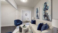 This well-appointed single-floor residence offers a cohesive blend of classic architectural character and modern finishes throughout its thoughtfully laid-out spaces. The living room serves as the heart of the home, featuring dark hardwood flooring, neutral walls, and curated décor accented with deep navy blue tones. Bold abstract artwork adorns the walls, complementing the elegant velvet seating and patterned area rug that anchor the space. An open doorway connects the living area to a separate dining room, where natural light filters in through large windows framed in dark trim, highlighting a radiator that speaks to the building's classic heritage. The kitchen is a standout feature, boasting rich, dark espresso cabinetry, white quartz countertops, a crisp subway tile backsplash, and stainless steel appliances including a gas range, all set atop light wood-tone flooring. Two bedrooms offer comfortable retreats; the primary bedroom is brightened by corner windows and features dark hardwood floors, while the second bedroom provides a serene, minimalist environment. The bathroom is elegantly finished with large-format grey floor tiles and striking marble-look wall tiles, paired with a grey shaker-style vanity, creating a clean and sophisticated atmosphere throughout.