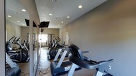 This fitness center offers a dedicated, single-floor space designed to support a comprehensive workout routine. The room is finished with neutral gray walls and durable tile flooring, creating a clean and functional environment. Recessed lighting is evenly distributed across the ceiling, ensuring the space is well-lit throughout.

Along one wall, large mirrors extend the visual depth of the room and are complemented by wall-mounted flat-screen televisions, allowing users to stay entertained during their sessions. A row of cardio machines lines the floor, including treadmills, ellipticals, and stationary bikes, providing a variety of aerobic exercise options.

Toward the far end of the room, a multi-station strength training rack is visible, complete with weight benches and cable systems to accommodate resistance training. A window near the strength equipment allows natural light to filter into the space, adding a welcoming element to the otherwise interior-focused room.

The layout is thoughtfully organized to allow ease of movement between equipment zones. The overall ambiance is practical and focused, catering to users seeking a straightforward, no-frills fitness facility. The space is well-maintained and presents all essential amenities needed for a complete and efficient workout experience.