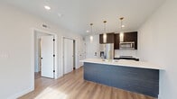 This thoughtfully designed single-floor residence showcases a contemporary aesthetic with high-quality finishes throughout. The open-concept kitchen serves as the centerpiece, anchored by a generous island clad in a blue-grey wood-tone finish and topped with a crisp white quartz countertop. A gold-tone faucet and undermount sink add refined detail, while three cylindrical glass pendant lights hang overhead, creating an elegant focal point. Dark espresso cabinetry lines the kitchen wall, housing stainless steel appliances including a french-door refrigerator, microwave, and range. Light hardwood flooring flows seamlessly throughout the entire unit, enhancing the bright, airy atmosphere created by white walls and recessed lighting. Bedrooms feature large floor-to-ceiling windows that flood the space with natural light and offer views of the surrounding urban streetscape, with one room providing direct access to a private balcony. Ample built-in closet storage is present in the bedroom areas. The bathroom is a standout feature, showcasing floor-to-ceiling marble-look tile in the walk-in glass-enclosed shower, complemented by a hexagonal mosaic tile floor, matte black fixtures, and a frameless glass door, delivering a spa-inspired finish.