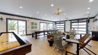 This single-floor amenity lounge is a thoughtfully designed social space that blends contemporary style with playful energy. The interior is anchored by light hardwood flooring, crisp white walls, and bold dark trim accents that frame the space with a modern industrial character. Recessed lighting throughout is complemented by a striking mid-century-inspired brass and globe chandelier that serves as a focal point above the central lounge area.

The space is well-appointed for recreation, featuring a full-size billiards table and a classic shuffleboard court. Multiple distinct seating arrangements invite both casual conversation and group gatherings, with a mix of woven wire chairs, slatted wood benches, and plush cushioned sofas accented with patterned throw pillows in green and black tones. A curved banquette runs along one wall, adjacent to a bold "ATL" letter installation set against a vertical wood slat accent wall, giving the space a strong sense of local identity.

Large black-framed windows and industrial-style glass garage doors flood the interior with natural light and offer views of the surrounding cityscape, including a connected outdoor terrace strung with bistro lights. A dining table with rattan chairs near the windows provides a casual spot to gather while enjoying the outdoor scenery. The overall ambiance is lively yet refined, making this lounge a versatile and inviting community hub.