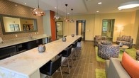 This single-floor space combines contemporary design with functional versatility, creating an inviting environment suited for gathering, work, or relaxation. The centerpiece of the room is a long, marble-topped communal table flanked by sleek black bar stools, ideal for group collaboration or casual dining. Above, a trio of copper pendant lights casts a warm glow, adding a refined industrial touch to the space.

The flooring throughout consists of rich, dark wood-toned tiles that ground the room and complement the warm yellow walls. An accent wall clad in exposed brick and painted in a bold terracotta orange introduces texture and visual interest, lending the space a modern-yet-approachable character.

Along one wall, a built-in kitchenette with dark cabinetry and a light countertop provides convenient access to a sink and additional counter space. Recessed lighting supplements the pendant fixtures, ensuring the room is well-lit throughout.

In an adjacent lounge area, colorful patterned armchairs are arranged atop vibrant green area rugs, creating a cozy retreat within the broader space. Natural light filters in through a window, enhancing the open, welcoming ambiance of the room.