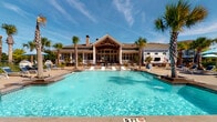 Located at 115 Great Lawn Dr in Summerville, SC, this single-story community amenity center offers a resort-inspired outdoor experience in the heart of the Lowcountry. The centerpiece is a sparkling resort-style swimming pool framed by a generous sun deck, complete with rows of lounge chairs and shade umbrellas for relaxed outdoor enjoyment. Decorative ceramic urn water features and black river rock accents add artful detail to the pool surround, while towering palm trees and a backdrop of lush pine forest create a serene, vacation-like atmosphere. A charming pergola structure anchors the far end of the pool area, enhancing the sense of a curated outdoor retreat. The building itself features classic coastal architecture with light blue siding, crisp white columns, and a dramatic exposed-timber gabled roofline. Herringbone-patterned brick flooring extends throughout the covered verandas, which are furnished with wicker seating, café-style tables, and ceiling fans for year-round comfort. Traditional gas lanterns and floor-to-ceiling French doors connect the interior seamlessly to the outdoor living spaces, flooding the interiors with natural light and offering direct views of the pool. An outdoor bar area with a mounted television provides additional entertainment options, making this amenity center a comprehensive gathering destination for the community.