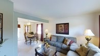 This two-floor residence offers a welcoming and functional interior highlighted by an open-concept main living area. The living room is generously proportioned, featuring light-toned carpet flooring, white walls, and a clean, simple ceiling that contributes to a bright and airy atmosphere. A large framed artwork adds a decorative focal point to the living space, while ample room accommodates a full sectional sofa and a glass-top coffee table, creating a comfortable area for relaxing or entertaining.

The living area flows seamlessly into the dining space, which is separated by a structural beam accent painted in a soft teal-green tone, adding a subtle architectural detail to the open layout. The dining area features a classic wooden dining set with seating for multiple guests, illuminated by a ceiling fan with integrated lighting. A window dressed with burgundy curtains allows natural light to filter into the space, enhancing its warm and inviting ambiance. The consistent carpet flooring throughout unifies both areas, contributing to a cohesive and comfortable living environment across the main level.