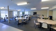 This single-floor communal space is designed to accommodate large groups in a functional, open layout. The room features multiple round tables paired with stackable chairs arranged throughout, providing flexible seating for dining, meetings, or social gatherings. The neutral-toned walls and dark carpeted flooring create a clean, understated aesthetic throughout.

Overhead, rows of fluorescent lighting ensure the space is consistently and evenly illuminated. Along one wall, large windows allow natural light to filter into the room, brightening the interior and offering views of the surrounding exterior. A wall-mounted flat-screen television is positioned at one end of the room atop a media console, adding a multimedia capability to the space.

Small decorative plants are placed on select tables, adding subtle touches of greenery throughout. The open floor plan allows for easy reconfiguration of the furniture to suit various event formats. The overall ambiance is practical and welcoming, making it well-suited for community dining, organized activities, or group presentations.