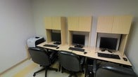 This single-floor space is designed as a dedicated computer lab, offering a clean and organized environment suited for focused work or study. The room features a row of individual workstations arranged along a shared light-wood laminate desk surface, each station equipped with a flat-screen monitor, keyboard, and mouse. Privacy panels in matching light wood extend upward from the desk, creating a sense of separation between users while maintaining an open and collaborative feel overhead. Each workstation is paired with a black ergonomic rolling chair, providing comfortable seating during extended use. A printer is conveniently positioned at one end of the workstation row, adding to the room's functional design. The space is finished with neutral-toned walls and light flooring, contributing to a calm and distraction-free atmosphere. Overhead cabinetry integrated into each workstation partition offers additional storage, keeping the workspace tidy and clutter-free. The overall layout is efficient and purpose-driven, making this space well-suited for academic, professional, or public computing needs.