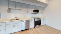 Located at 8585 La Mesa Blvd in La Mesa, San Diego, this single-floor unit offers a clean, modern aesthetic defined by light wood-style flooring, crisp white walls, and recessed lighting throughout. The open-concept living and kitchen area is anchored by a well-appointed kitchen featuring soft gray shaker-style cabinetry, light quartz countertops, and a full suite of stainless steel appliances including a refrigerator, electric range, over-the-range microwave, and dishwasher. An in-unit stacked washer and dryer is conveniently tucked into a dedicated laundry area just off the kitchen. The living area is bright and open, with recessed lighting and direct access to a front entry door flanked by a large window that invites natural light and views of the surrounding outdoor landscaping. The bedroom is generously sized, featuring a ceiling fan, multiple windows offering outdoor views, and a full-width mirrored sliding closet that enhances the sense of space. The bathroom is well-designed with a tub-shower combination, white tile surround, built-in shelving, a single-sink vanity with a light countertop, and matching light wood-style flooring. The overall ambiance of the space is bright, clean, and move-in ready, with a cohesive modern design palette throughout.