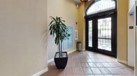 The South Lobby serves as a primary entry point, combining functional amenities with an inviting architectural character. The space is finished with warm terracotta ceramic tile flooring laid in a diagonal pattern, which extends throughout and contributes to a cohesive, Mediterranean-inspired aesthetic. Notably tall ceilings lend the lobby a sense of openness and volume, amplified by the warm beige textured walls.

The main entry features a set of elegant double doors framed in dark metal with ornate ironwork detailing and a large arched transom window above, allowing natural light to filter into the space. A decorative pendant fixture is visible overhead, adding to the ambient warmth of the entry. A tall, large-leafed indoor plant in a square black planter provides a striking natural focal point near the entrance.

Along the interior wall, a bank of individual resident mailboxes offers convenient mail access. A fire extinguisher cabinet and fire alarm are mounted nearby, ensuring safety compliance. An additional interior door with a louvered vent panel leads to a service or utility room. The overall ambiance is clean, functional, and welcoming, reflecting a well-maintained shared entry environment.