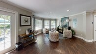 Located at 1904 US-80 in Garden City, Georgia, this single-story space is thoughtfully designed to offer both functional and welcoming environments. The central reception area commands immediate attention with its dramatically raised tray ceiling, clerestory windows that draw in abundant natural light, and a striking multi-pendant chandelier with amber-toned shades. A large, light-finished reception counter with bar-height seating anchors the space, flanked by decorative white columns that define zones while maintaining an open, airy feel throughout.

Rich, dark hardwood flooring flows seamlessly across the entire space, complementing the neutral warm-beige wall tones and crisp white crown molding and baseboards. Recessed lighting is used throughout, enhancing the brightness provided by multiple windows fitted with plantation shutters and patterned drapery panels.

The lounge area features a curated seating arrangement with tufted accent chairs, a cushioned ottoman, and a decorative console adorned with artwork and table lamps. A beverage station is conveniently positioned nearby. A separate dining or meeting nook is visible toward the rear, complete with round tables and upholstered chairs. French doors and multiple entry points offer easy access, contributing to the space's open, connected ambiance.