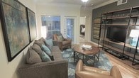This thoughtfully designed single-floor space showcases a harmonious blend of contemporary and transitional style. The living area serves as the heart of the home, featuring elegant crown molding and warm hardwood-style flooring that adds a sense of refinement throughout. Natural light filters generously through multiple windows adorned with horizontal blinds, brightening the space and creating an inviting atmosphere. A sliding glass door leads to an outdoor area, further enhancing the sense of openness.

The living room is anchored by a plush sectional sofa accented with soft teal and blue throw pillows, complemented by a classic arm chair and a rich leather accent chair. A round wooden coffee table sits atop a decorative area rug with a medallion pattern, tying the seating arrangement together cohesively. An industrial-style metal shelving unit mounted against a dark feature accent wall adds a bold, modern contrast to the otherwise neutral color palette.

A glimpse into the adjacent bedroom reveals a neatly made bed, suggesting a seamless and functional flow between living spaces. The overall ambiance is warm, curated, and comfortable, making this space well-suited for both everyday living and entertaining.