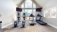 Located at 422 Charleston Green in Malvern, PA, this single-story fitness center is defined by its striking architectural design, most notably a dramatic vaulted ceiling that rises to a prominent peak. A large, multi-pane arched window arrangement at the front of the space floods the interior with natural light and offers open views of the surrounding neighborhood, creating an airy and energizing workout environment. Additional windows along the side walls further enhance brightness throughout the day.

The fitness center is comprehensively outfitted with a wide selection of cardio and strength training equipment. Cardio offerings include multiple treadmills, elliptical machines, a StairMaster, and a rowing machine, accommodating a variety of aerobic workouts. For strength training, the space features a Smith machine, a cable machine system, a dumbbell rack with a full range of weights, and additional free weight equipment. Wall-mounted flat-screen televisions are positioned throughout, providing entertainment during workouts.

The flooring consists of durable, neutral-toned carpet tiles laid in a herringbone pattern, adding both comfort underfoot and visual appeal. The overall ambiance is bright, open, and well-organized, with neutral-toned walls complementing the spacious layout and making efficient use of the room's generous footprint beneath its soaring roofline.