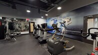 Located at 9901 Washington Blvd in Los Angeles, this modern fitness center is designed to inspire and accommodate a full range of training needs. The single-floor facility features a dark, industrial aesthetic with exposed ductwork along the ceiling, recessed and pendant lighting that evenly illuminates the workout floor, and durable rubber flooring throughout. A striking large-scale athletic mural dominates one wall, depicting a sprinter in motion against a cityscape backdrop, setting an energetic and motivational tone for the space. The cardio zone is equipped with a row of Octane ellipticals and Life Fitness treadmills, providing ample options for cardiovascular training. Adjacent to this area, a strength training section houses a Life Fitness cable and pulley machine system, a functional training bench, and a well-stocked dumbbell rack with medicine balls and stability balls. Full-length mirrors line one wall, enhancing the sense of space and allowing users to monitor their form. Battle ropes, resistance bands, and a spin bike round out the functional fitness offerings. Restrooms are conveniently located within the facility. The overall ambiance is sleek, professional, and purpose-built for performance.