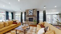 Located at 1300 South Park Way in Sanford, NC, this single-story community clubhouse offers a well-appointed gathering space designed for both relaxation and productivity. The main lounge is a standout area, anchored by a dramatic floor-to-ceiling stacked stone fireplace with a rustic wood mantel. Vaulted ceilings, recessed lighting, and rich hardwood flooring in a herringbone pattern contribute to the room's warm and inviting ambiance, while large windows and French doors flood the space with natural light and provide views of the outdoor pool area. The open-concept kitchen features rich mahogany cabinetry, dark granite countertops, stainless steel appliances, a central island with bar seating, and pendant lighting overhead — ideal for community events and gatherings. Adjacent to the kitchen, a casual dining area with a round wood table and leather chairs sits beside glass French doors that open to the pool courtyard. A separate game room is visible in the floor plan, offering additional recreational space. The clubhouse also includes a dedicated business center equipped with desktop computers, a printer, and granite work surfaces, providing residents with convenient access to productivity tools. Decorative wainscoting and architectural columns add refined detail throughout the space.