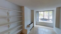This single-floor space presents a clean, minimalist aesthetic defined by neutral tones and functional design elements. The main living area features light hardwood flooring that adds warmth to the otherwise crisp white interior. A prominent built-in shelving unit lines one wall, offering generous vertical storage across multiple open shelves — a practical and architectural feature that draws the eye. Adjacent to the shelving, a low-profile baseboard heating unit sits flush against the wall, maintaining the streamlined look of the space. The large picture window on the far wall serves as a focal point, flooding the room with natural light and framing views of the surrounding streetscape and greenery. The textured ceiling adds subtle visual interest overhead. Overall, the space conveys a sense of openness and simplicity, well-suited for those who appreciate an uncluttered, functional environment with thoughtful built-in storage solutions.