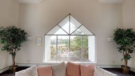 This single-floor space makes an immediate architectural statement through its defining feature: a striking floor-to-peak gabled window wall in the living area. The triangular glazed structure mirrors the roofline's pitch, drawing the eye upward to soaring vaulted ceilings finished in crisp white. The expansive window composition—framed by clean metal mullions—allows an abundance of natural light to pour into the interior throughout the day, creating a bright and airy atmosphere. Looking outward through the glass, the view reveals a picturesque landscape of mature trees and open greenery, with an ornamental balcony railing visible just beyond the window, adding a decorative architectural layer to the outdoor setting. The living area itself features soft, neutral wall tones that complement the natural palette of the views beyond. A generously sized sofa dressed in muted blush and ivory cushions anchors the seating area beneath the window, reinforcing the space's calm and welcoming character. The overall ambiance is one of openness and tranquility, where thoughtful architectural design and generous daylighting work together to create a comfortable, light-filled environment that feels connected to the natural surroundings outside.